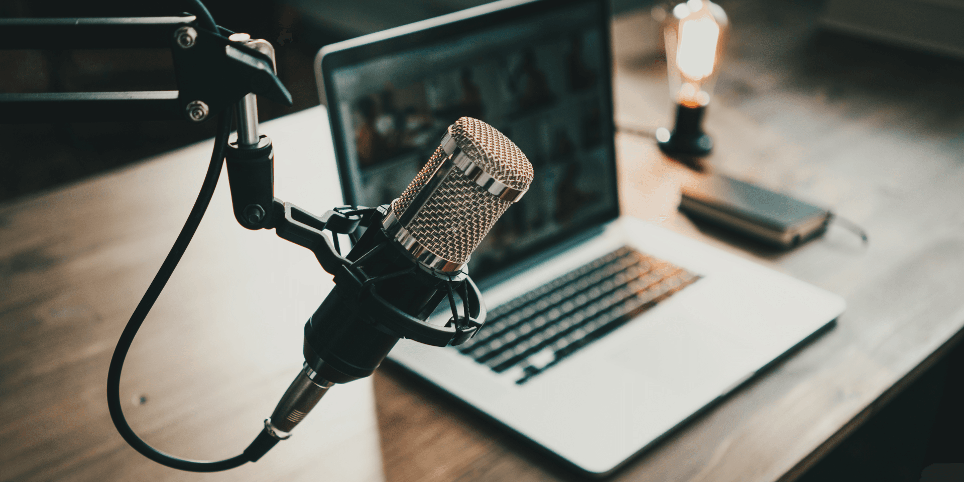 View of a laptop and professional podcast microphone on a wooden desk with a warm light in the background